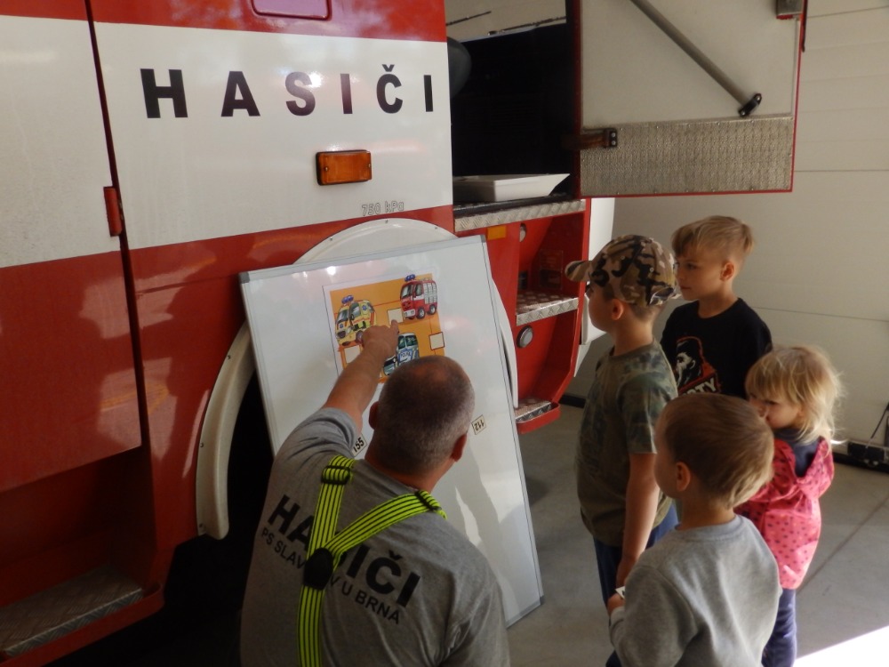 Děti stojí při hasičském autě a poslouchají hasiče, který jim něco ukazuje