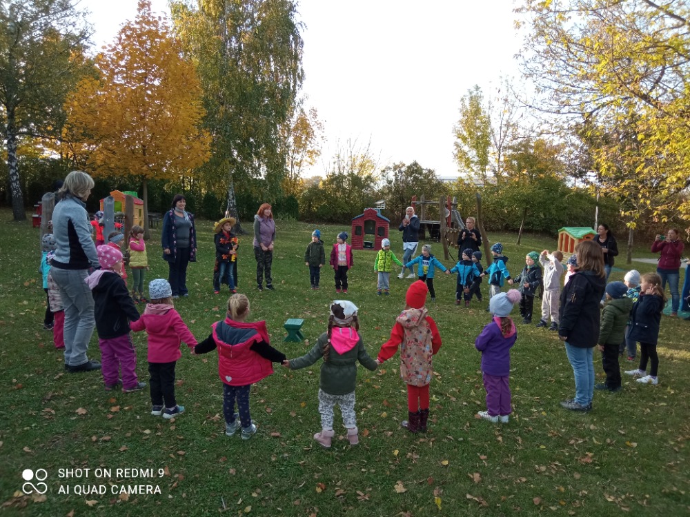 Děti stojí v kruhu na zahradě a zpívají